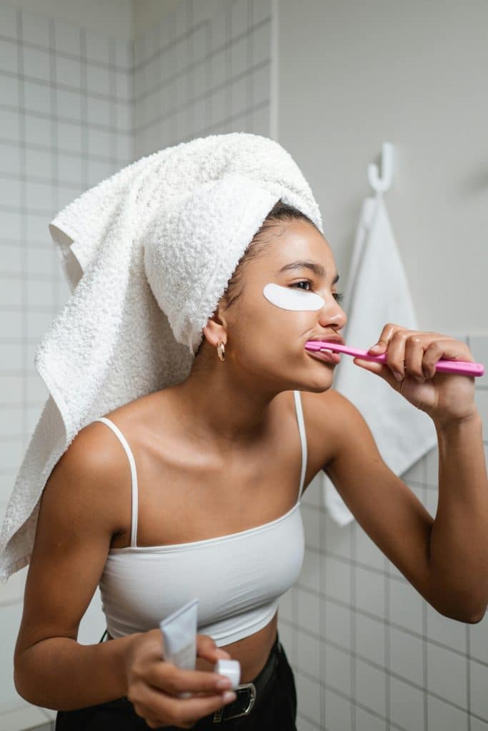 Woman with a towel on her head brushing teeth, wearing under-eye patches and a white tank top.