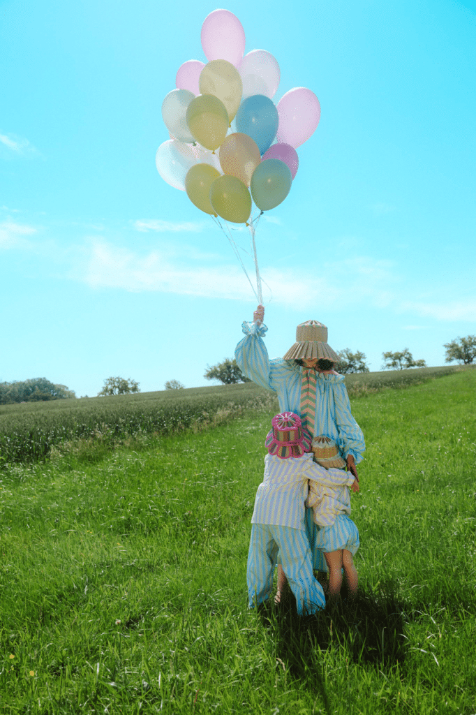 Adult and two children in striped pastel outfits and straw hats stand in a grassy field holding colorful balloons.