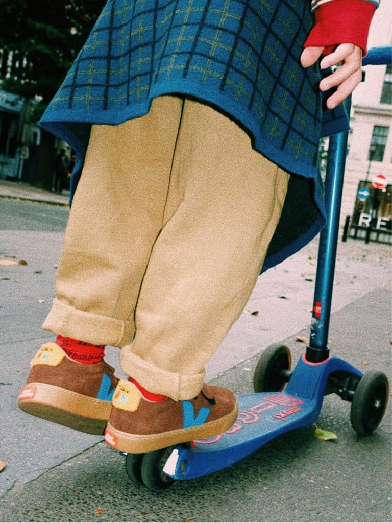 Child wearing brown VEJA x Jelly Mallow trainers with blue V logos, beige trousers and a blue checked top while riding a scooter.