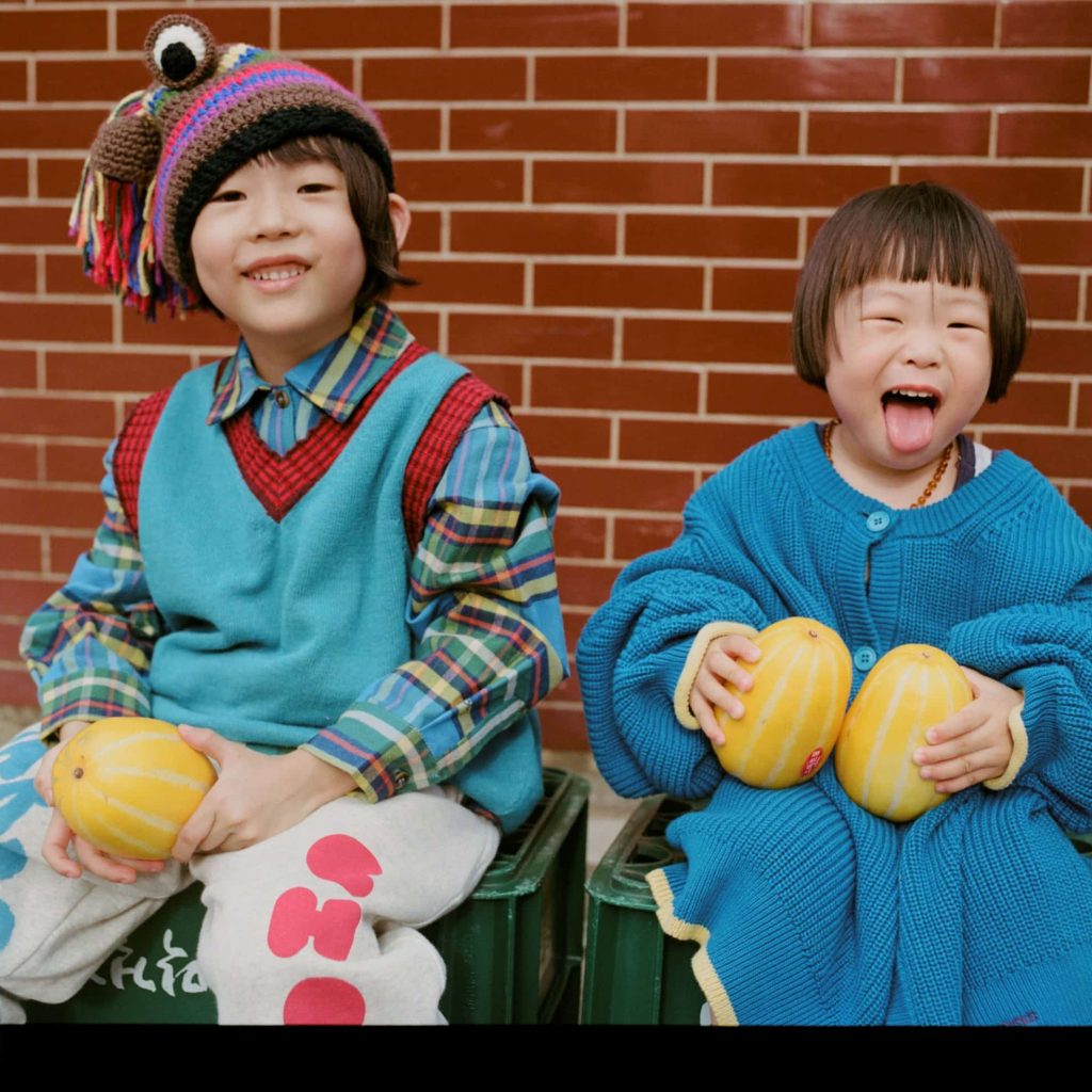 Two children sit on green crates against a brick wall, smiling and holding yellow melons, wearing teal sweaters.