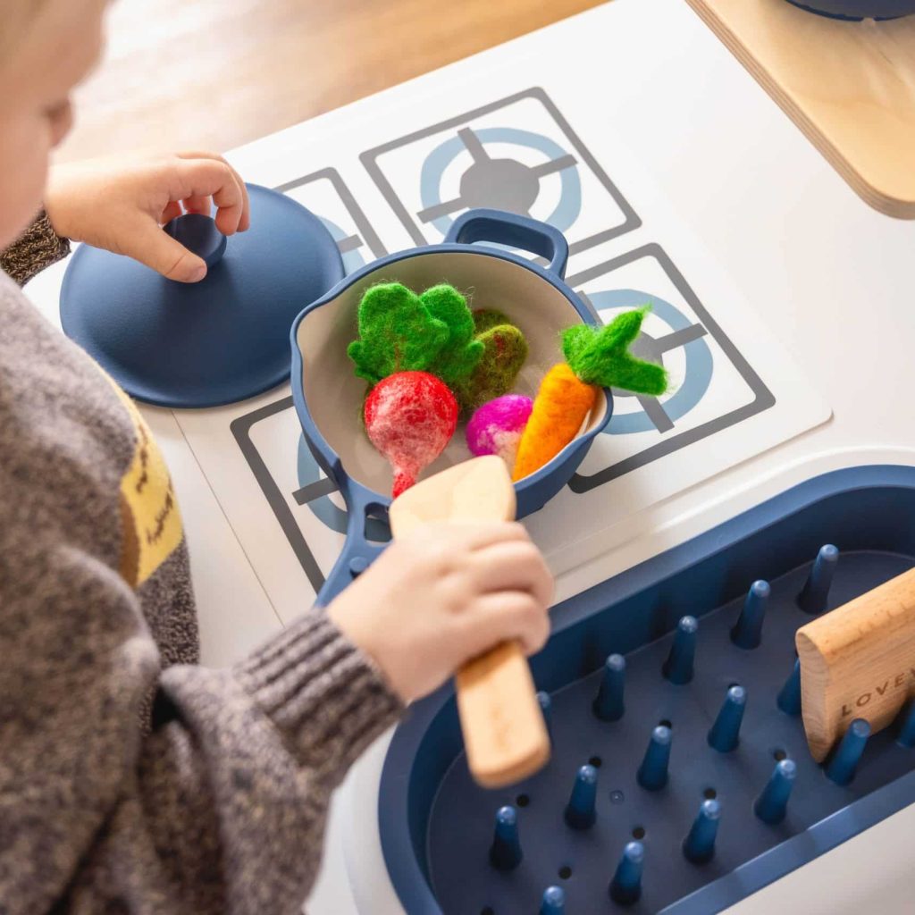 Child in a grey sweater playing with a toy kitchen, stirring felt vegetables in a pot on a play stove.